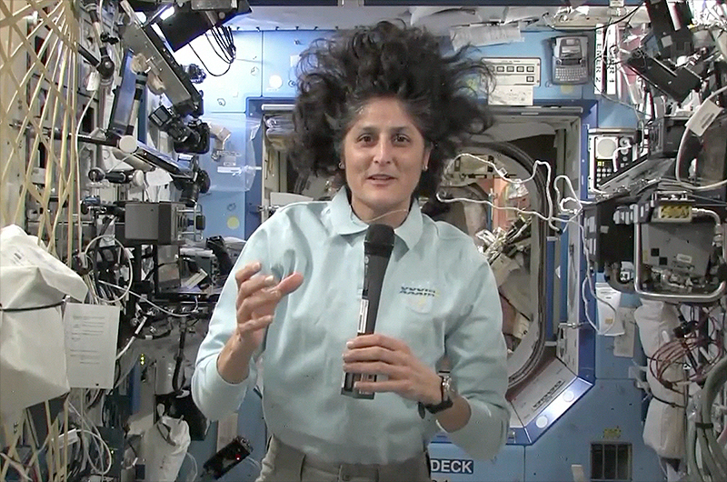 NASA astronaut Sunita Williams floating in microgravity inside the International Space Station while holding a microphone and gesturing during a broadcast.