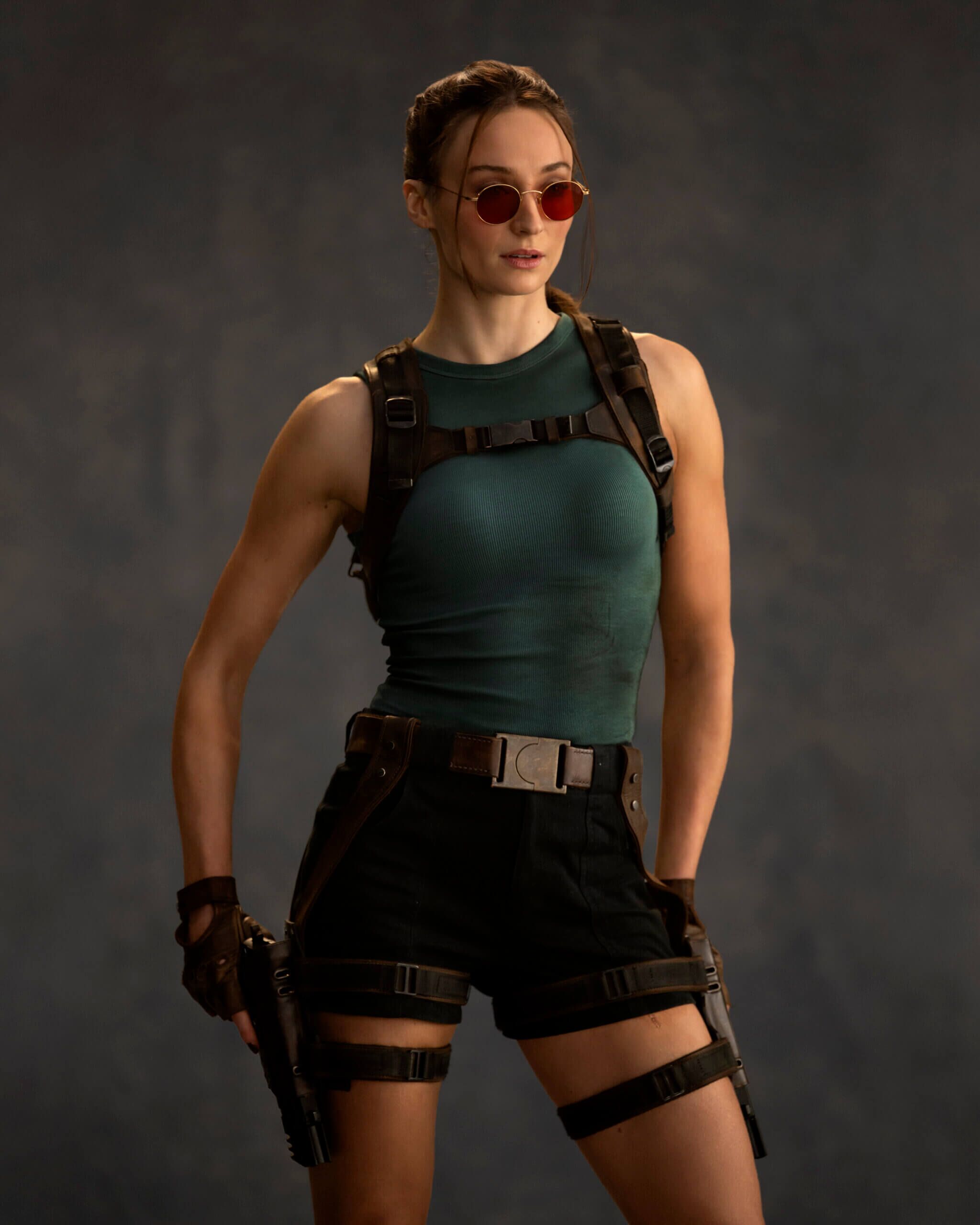 Sophie Turner as Lara Croft in a wardrobe test for Prime Video's Tomb Raider, wearing a teal tank top, brown shorts, and signature red sunglasses.